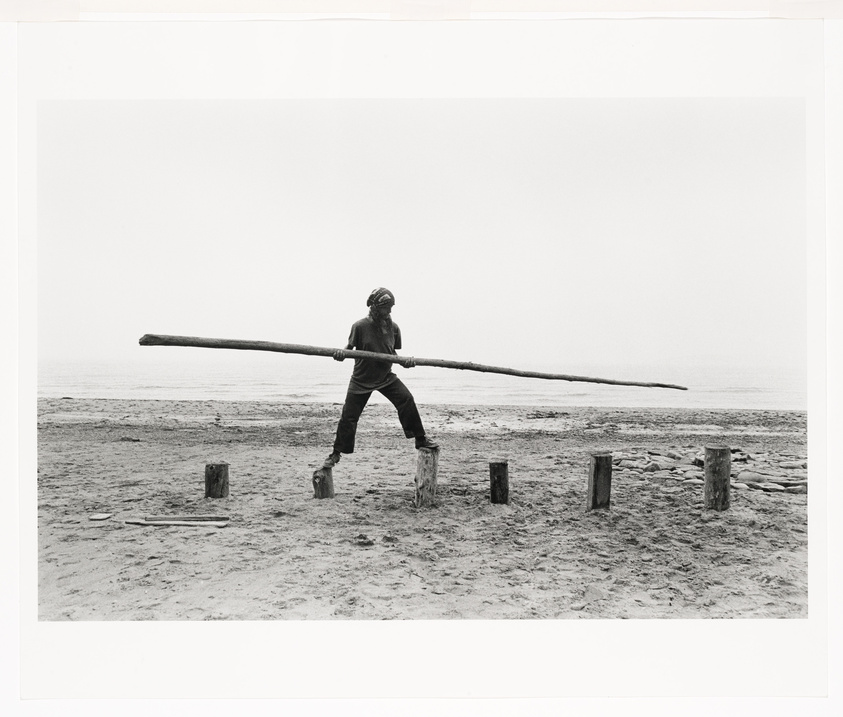 Person balancing on wooden posts at the beach, holding a long pole horizontally.