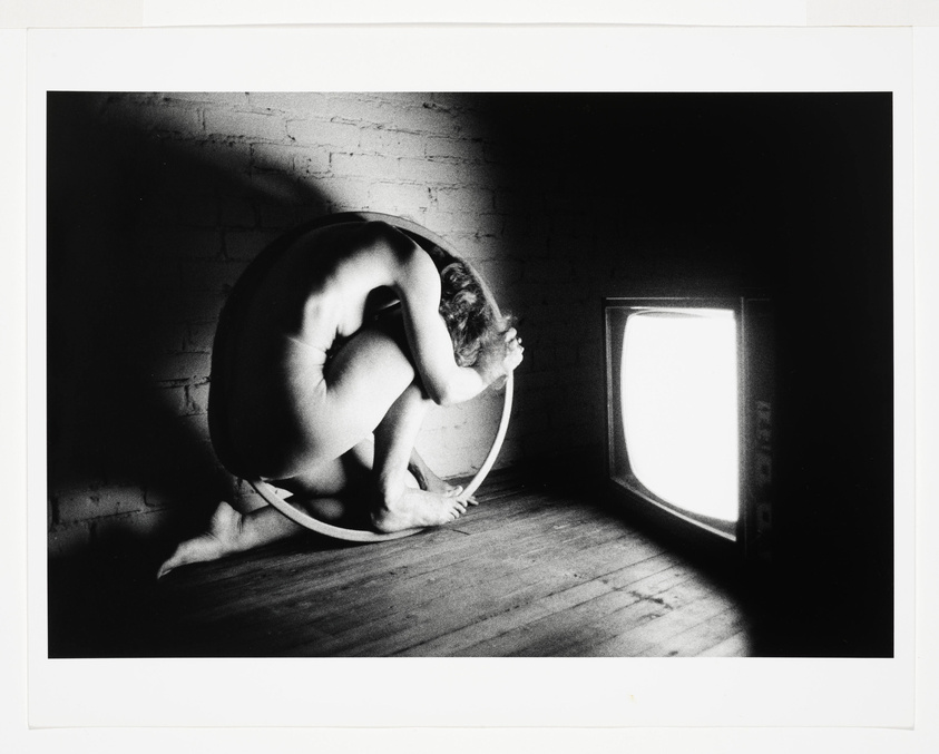 A black and white artistic photograph depicting a nude person curled up inside a large hoop, with their head resting on the hoop's lower curve. The person is positioned in a dimly lit room with a brick wall and wooden floor, near a glowing television set that is casting light on the scene. The image conveys a sense of solitude and introspection.