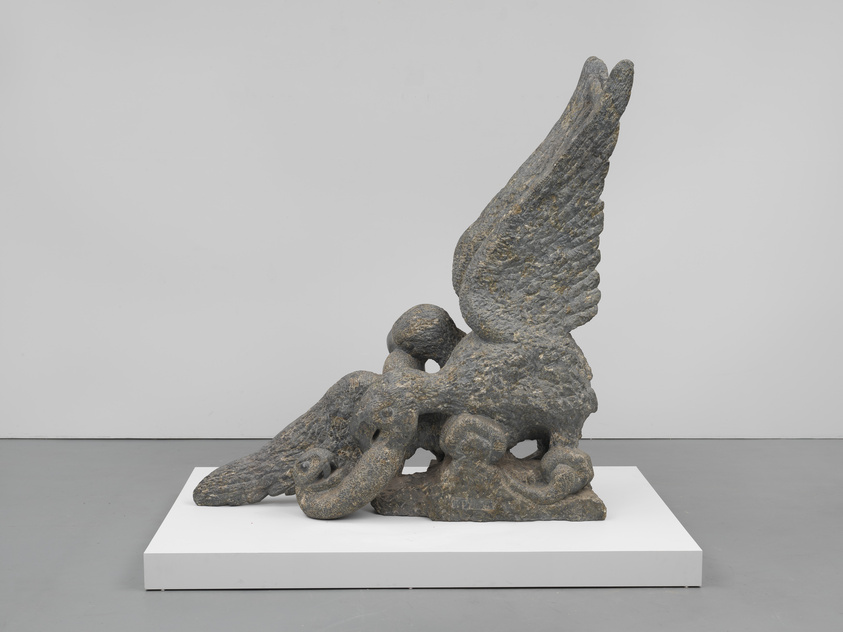 A stone sculpture of a winged creature, possibly a mythical being or an angel, is displayed on a white pedestal against a plain background. The figure has one large, detailed wing extended upwards, while the other is not visible, suggesting it might be broken or tucked away. The creature's head is bowed down in a contemplative or sorrowful pose.