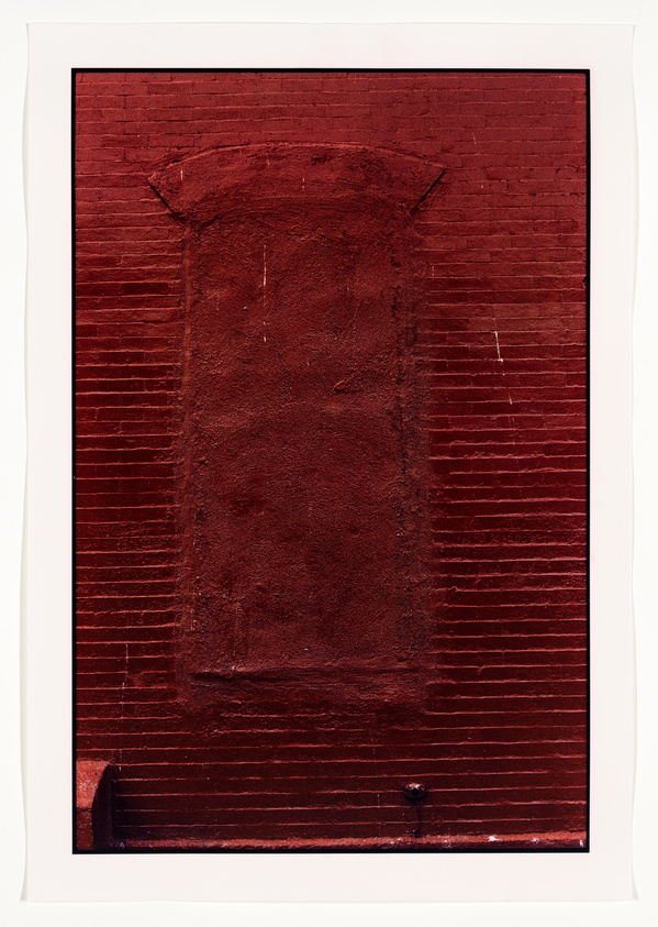 A photograph of a red-painted brick wall with a sealed-up doorway that has been covered over with bricks, creating a textured rectangular outline where the door once was. A single black doorknob remains on the wall, adding a contrasting detail to the scene. The image is framed with a white border.