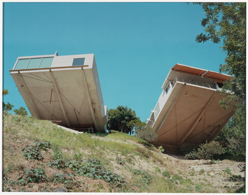 Two unique, boat-shaped houses perched on a hillside with their pointed ends angled sharply towards the sky, surrounded by greenery under a clear blue sky.