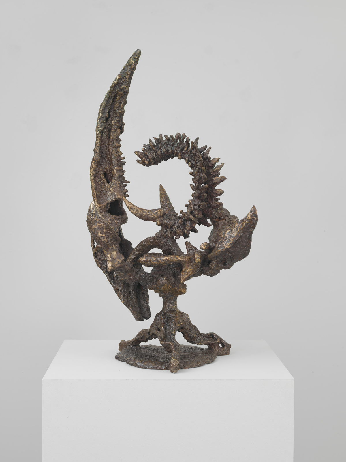 A bronze sculpture with abstract and organic forms is displayed on a white pedestal against a light background. The sculpture features textured surfaces and appears to be composed of intertwining elements with some parts resembling natural shapes like branches or bones.
