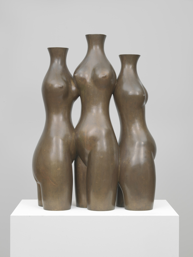 A sculpture of three abstract female figures merged at the torso, with simplified forms and no arms, heads, or legs, displayed on a white pedestal against a light background.