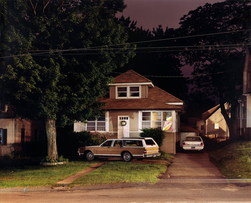 A nighttime scene of a residential street with two-story houses, illuminated by streetlights. A large tree dominates the left side of the frame. In the foreground, an older model station wagon is parked on the street in front of a house with a light brown exterior and white trim. A wreath hangs on the front door. To the right, another house with a lit window displays a neon "Open" sign. Power lines stretch across the dark sky above.