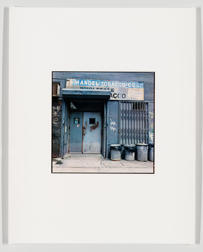 A photograph of an old, weathered storefront with a sign reading "Mandel Tobacco Co. Inc." above a closed, graffiti-tagged metal gate and a door with peeling blue paint. Three trash cans are lined up against the wall to the right of the door. The image is mounted on a white background with a wide border.
