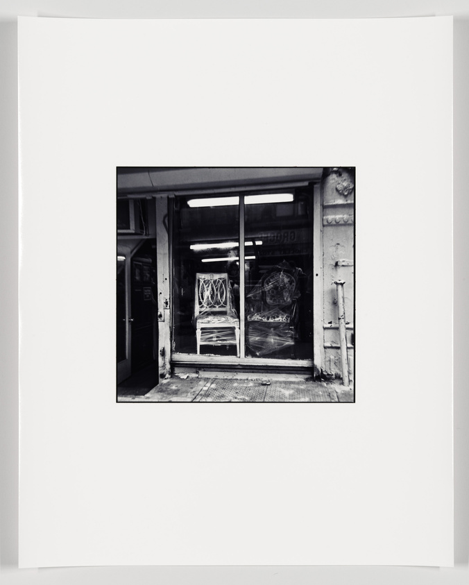 A black and white photograph of a framed artwork depicting a weathered storefront with graffiti on the glass doors.