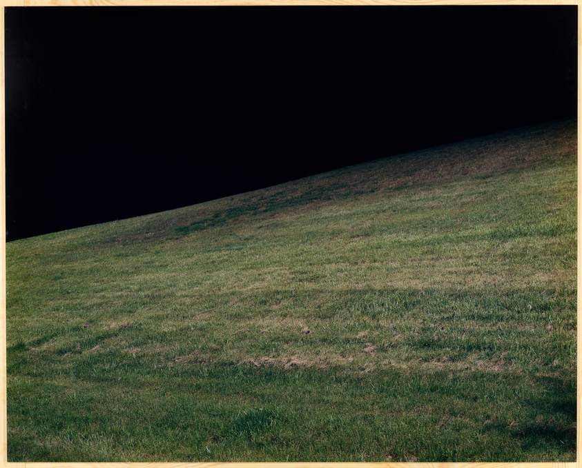 A grassy hill slopes upward from the bottom right corner to the upper left corner against a pitch-black background, creating a stark contrast between the dark sky and the illuminated grass.