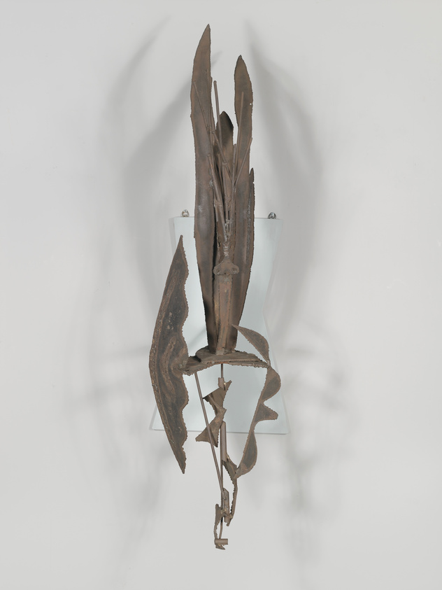 A vertical metal sculpture resembling abstract plant forms or feathers, mounted on a white wall with shadows casting an intricate pattern behind it.