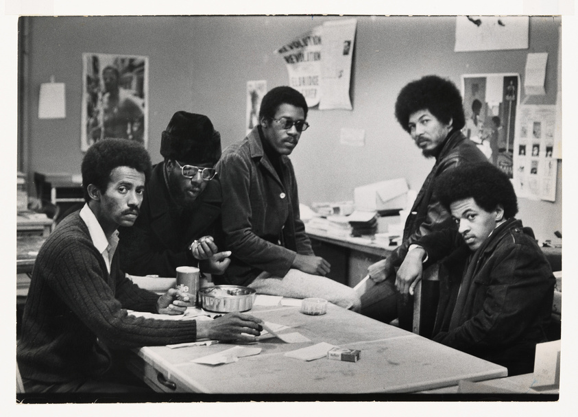 Five men gathered around a table in a room with activist posters, looking intently at the camera.