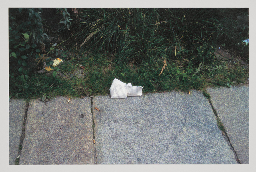 A discarded crumpled white paper bag lies on a gray sidewalk next to overgrown green grass with a few scattered cigarette butts and a small yellow packet nearby.