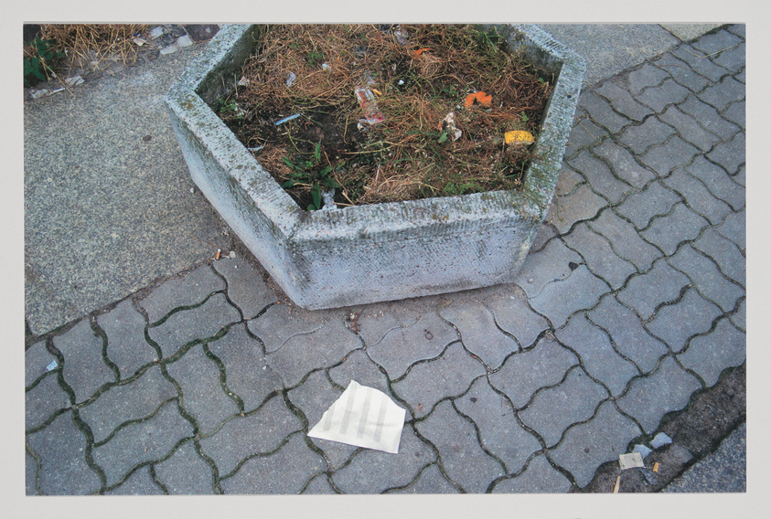 A weathered concrete planter filled with dry plants and litter on a paved sidewalk, with a discarded newspaper lying on the ground nearby.