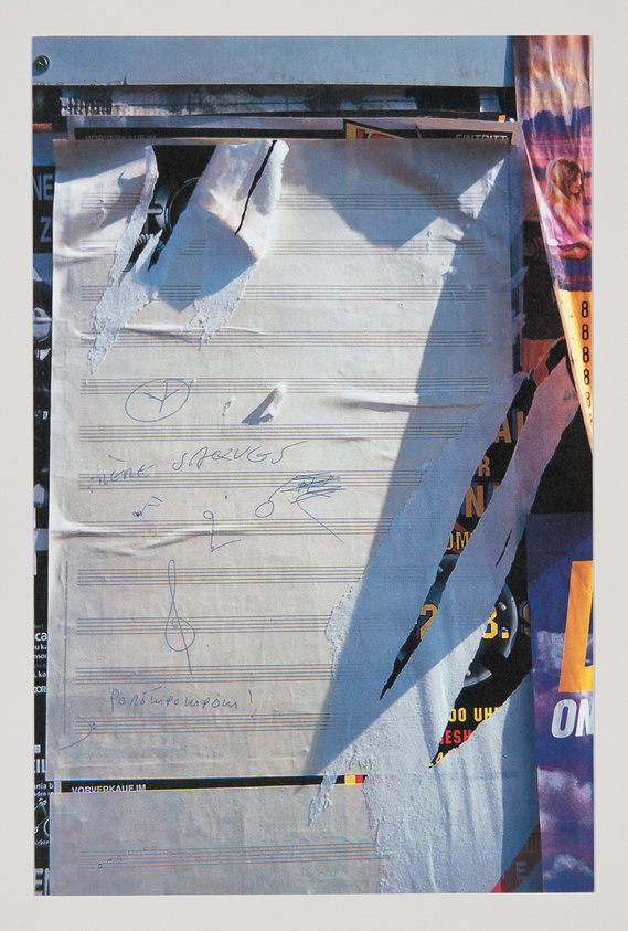 A close-up photograph of a torn poster wall with various layers of paper and advertisements. The visible top layer features musical staff lines with several signatures and scribbles. The torn sections reveal underlying layers of colorful posters with text and images partially visible.