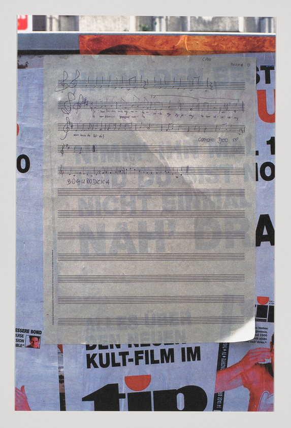 A weathered sheet of music paper with handwritten notes and lyrics is taped to a wall covered with partially visible posters, including one with text in German and another featuring a small image of James Bond. The music sheet has annotations and doodles, including a heart and the words "cantiche d'Iddio."