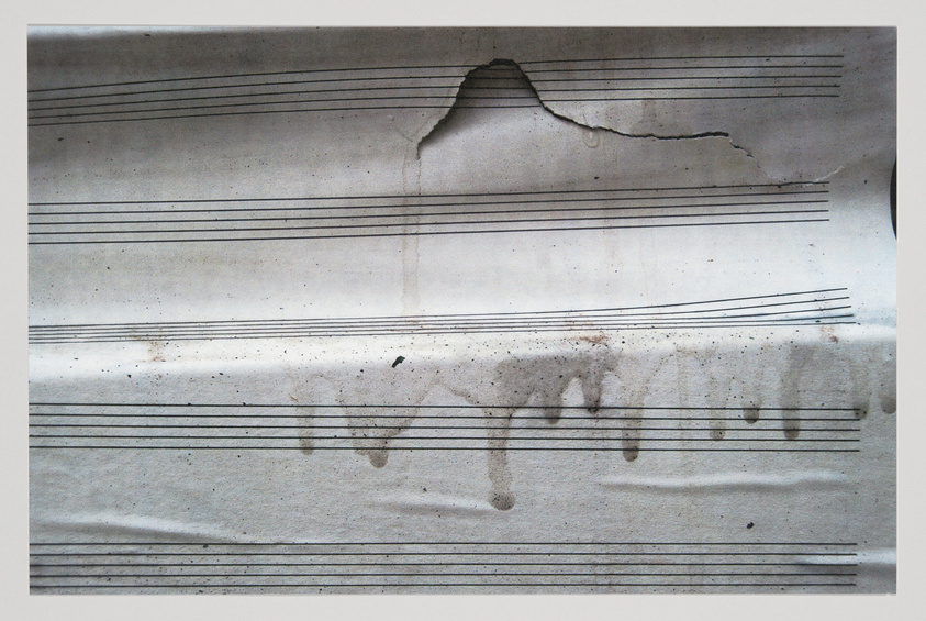 A worn piece of sheet music with blank staves, showing signs of water damage and a large tear at the top center, against a white background.