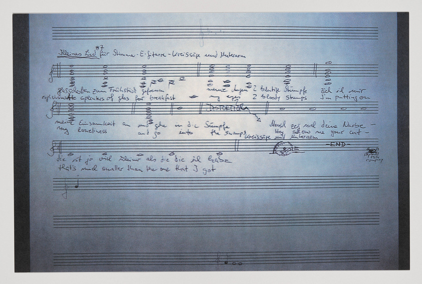 A photograph of a handwritten sheet of music with lyrics and chord annotations. The page shows signs of editing and personal notes, indicating a work in progress. The background is a gradient from white to blue, suggesting the sheet may be on a blue surface or affected by a lighting effect.