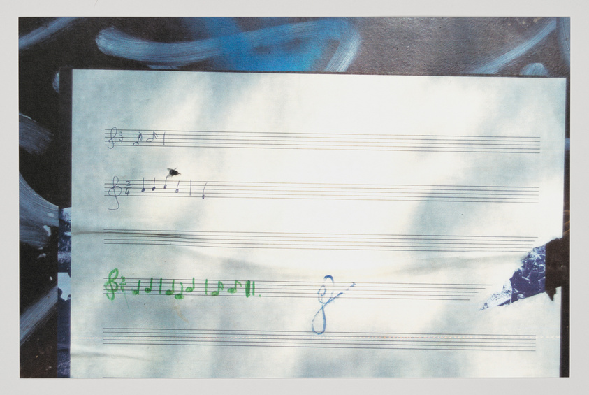 A photograph of a sheet of music paper with a few musical notes and symbols drawn in different colors, overlaid on a background with abstract blue and black brushstrokes. The paper appears worn and slightly crumpled.