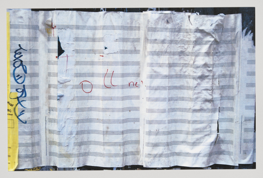 A worn and creased piece of paper with musical staff lines printed on it, showing various marks, stains, and doodles, including a large blue scribble on the left and red markings that resemble a circled number and a word fragment. The paper appears to be taped or pinned against a wall or board.