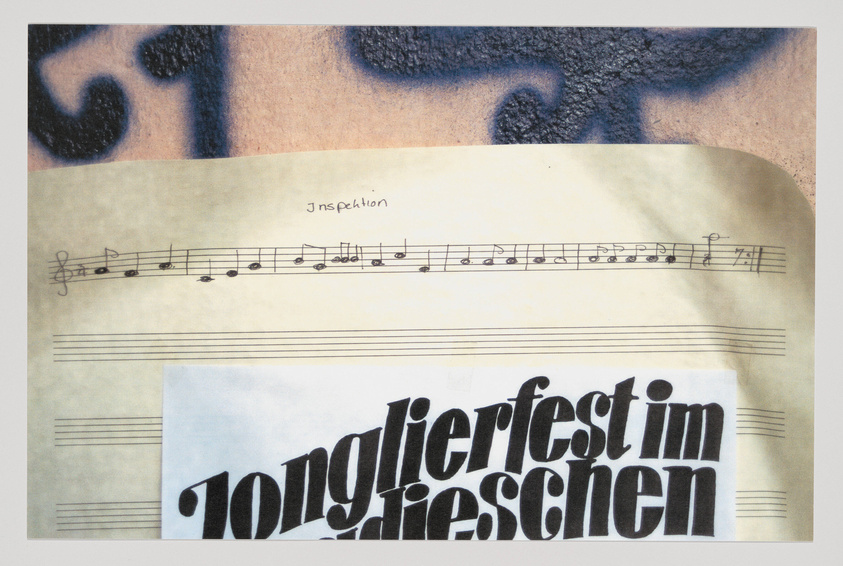 A photograph of a piece of paper with musical notation on it, placed over a printed text that reads "jonglierfest im rieschen." The paper with music notes appears aged and is partially covering another layer with a large, blurred tattoo-like design in the background.