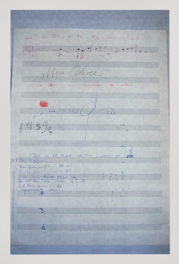 A photograph of a sheet of paper with various handwritten notes, musical notations, and scribbles in different colors of ink. Some text is legible, including phrases like "This is no place for the luxury of love," while other parts are in cursive or another language, making them harder to read. There are also some red ink stamps and blue pen markings scattered across the page.