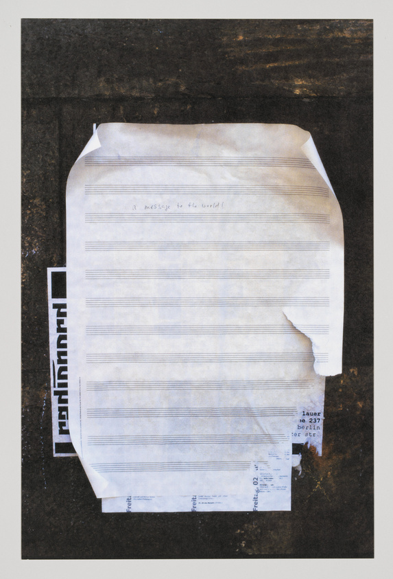A crumpled piece of lined paper with the handwritten phrase "a message to the world" is taped to a dark, textured background alongside a torn label with an address in Berlin.