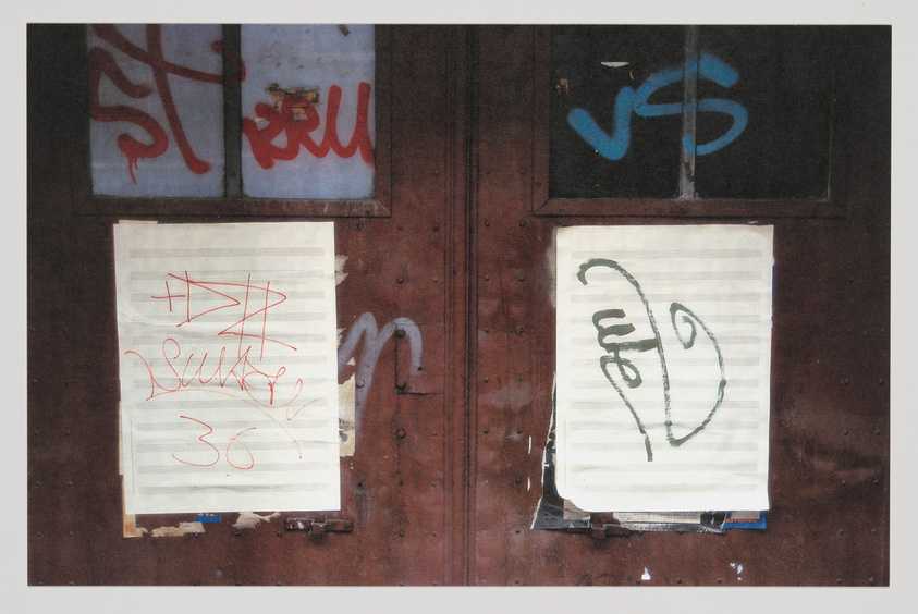 A weathered double door with graffiti and two pieces of paper taped over the windows. The left paper has red scribbles and the number 36, while the right paper has a black scribble resembling a heart. The doors are adorned with rust and peeling paint, suggesting abandonment or disrepair.