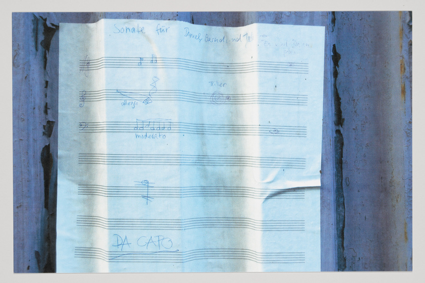 A crumpled piece of paper with handwritten musical notations and annotations pinned to a wooden surface with visible signs of weathering. The text on the paper includes musical directions such as "allegro" and "moderato," as well as the phrase "DA CAPO." There are also personal notes written at the top of the sheet.