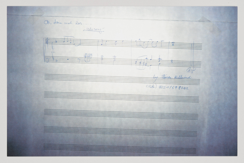 A photograph of a sheet of music paper with a handwritten musical score at the top. The title "Oh, dann mal los..." is written above the staff, and "Ideenskizze" (sketch of ideas) is noted beside it. The composer's name, Martin Behrmann, is written at the end of the musical notation, along with a date, possibly "02/21/1982". The rest of the sheet is blank, with empty staves ready for additional music notation.