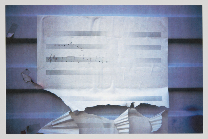 A crumpled sheet of music paper with several musical notes and symbols printed on it, displayed against a blurred background with blue hues. The paper is torn in several places, creating a sense of neglect or abandonment.