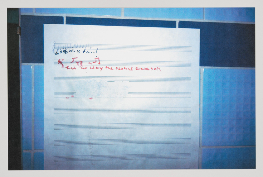 A photograph of a piece of paper with musical staff lines taped to a tiled wall. The paper has handwritten musical notes and red annotations, including some text that appears to be corrections or instructions related to the music. The image has a bluish tint, suggesting it may have been taken in low light or with a filter.