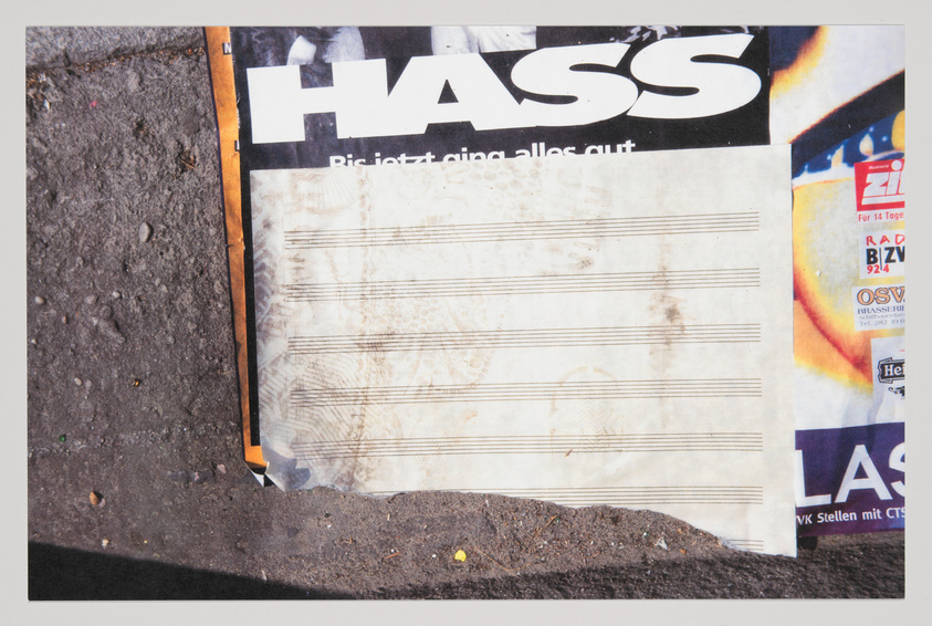 A torn and weathered poster on a wall with the word "HASS" in large black letters at the top, followed by smaller text that reads "Riskiert, alles aus." Below this headline is a blank area with faded horizontal lines resembling a musical staff. The poster is partially overlapped by other layers of torn posters and is set against a gritty, textured background.