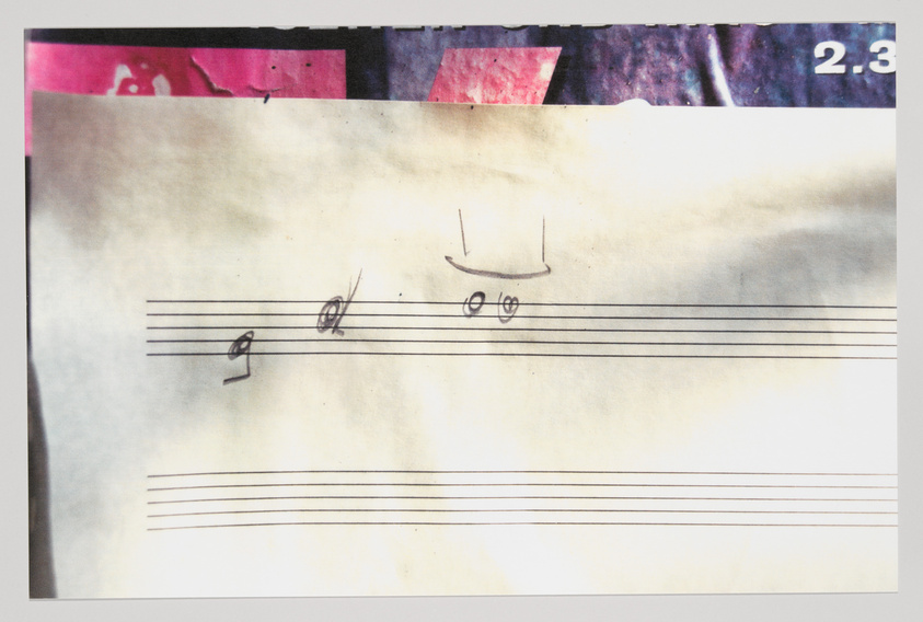 A photograph of a piece of paper with musical staff lines and a few hand-drawn music notes, showing signs of wear and age. The top of the image has a strip of another image with pink and purple hues and the numbers "2.3".