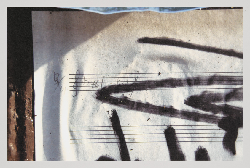 A close-up photograph of a piece of paper with musical staff lines and a few notes, overlaid with a large, abstract black graffiti mark. The paper appears aged and slightly crumpled, with the graffiti giving a sense of urban art intervention on a musical composition.