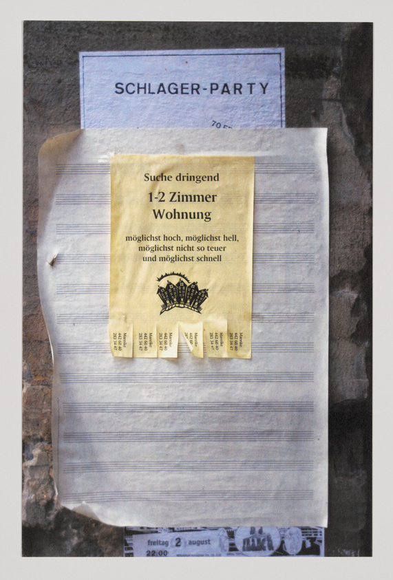 A photograph of a bulletin board with multiple layers of flyers, the topmost being a yellow flyer in German seeking urgently a 1-2 room apartment, with tear-off contact tabs at the bottom. Behind it, partially visible, is a blue flyer for a 'Schlager-Party' and another flyer with musical notation lines. The board appears weathered, and the flyers show signs of wear and tear.