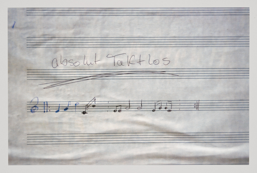 A photograph of a piece of paper with musical staff lines and handwritten musical notes. Above the staff, the words "Absolut Tarklos" are written in cursive. The paper appears slightly wrinkled.
