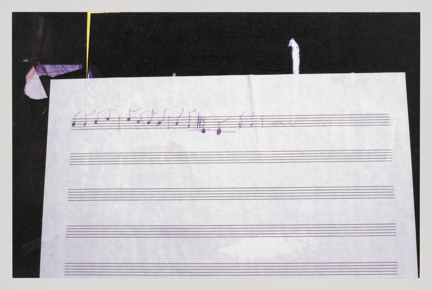 A piece of paper with musical notation is partially visible, with handwritten notes on a staff, against a dark background with torn edges.