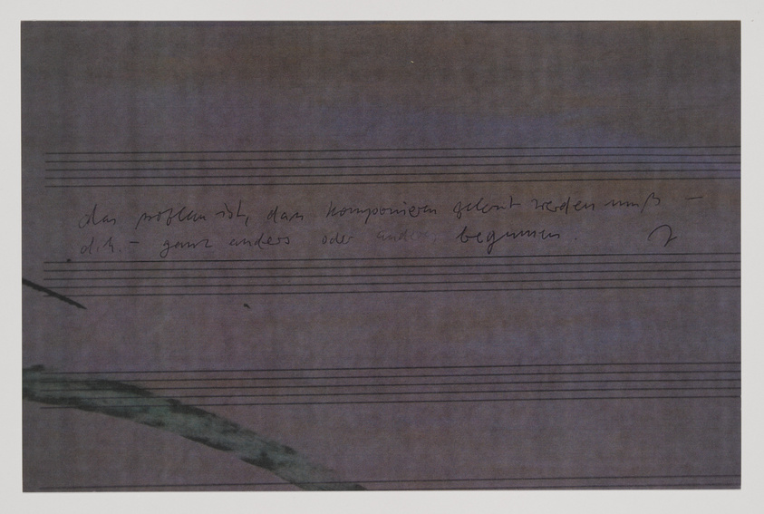 A photograph of a piece of paper with musical staff lines and handwritten German text, with a dark, mottled background and a faint greenish brushstroke on the bottom left side.