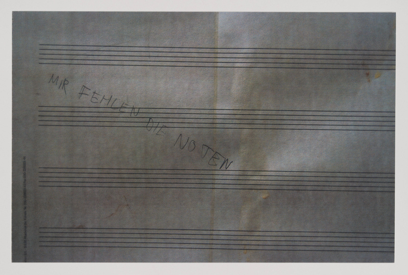 A photograph of a piece of paper with musical staff lines and a handwritten German phrase "MIR FEHLEN DIE NOTEN" which translates to "I am missing the notes." The paper appears aged with some discoloration and spots.