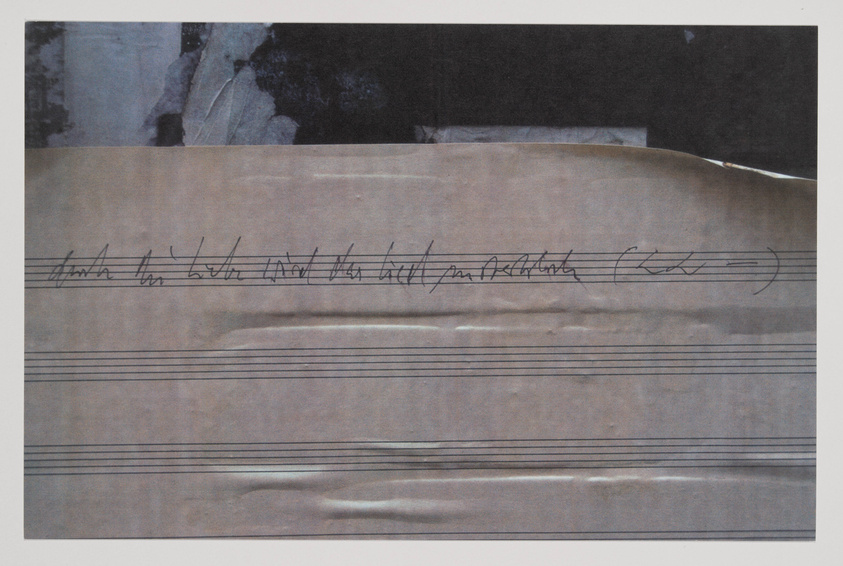 A worn piece of sheet music with handwritten musical notation and scribbles, showing signs of aging and use, with a dark, indistinct background at the top of the image.