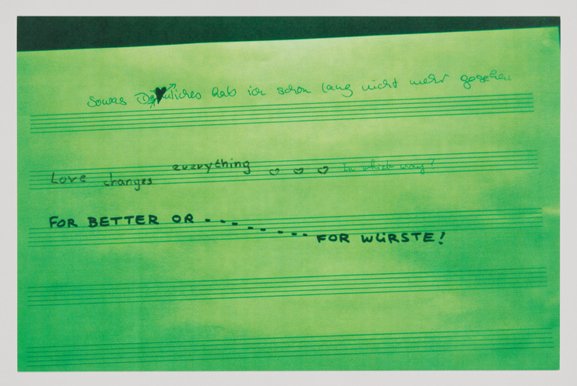 A photograph of a green-tinted paper with handwritten text and music staff lines. The text includes phrases like "Love changes everything" and "FOR BETTER OR ... FOR WÜRSTE!" with some words crossed out and hearts drawn above the staff lines.