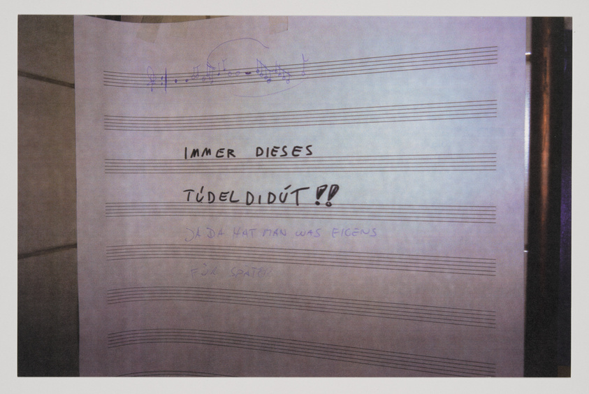 A photograph of a piece of paper with musical staff lines and handwritten notes in German, including the phrase "IMMER DIESES TÜDELIDÜT!!" with some musical notes and additional text scribbled below. The paper appears to be taped to a wall or a surface with a reflective edge on the right side.