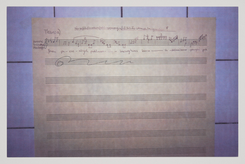 A photograph of a sheet of music paper with handwritten musical notation and annotations. The paper is displayed against a background with blue and purple hues.