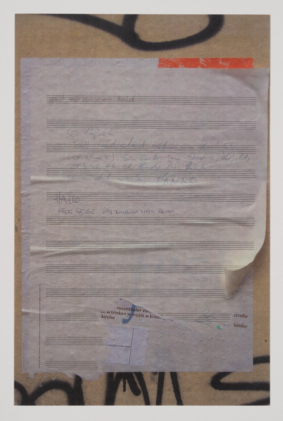 A collection of weathered papers with handwritten notes and printed text is taped to a wall with graffiti. The papers appear to be partially covering each other, and some text is obscured. The visible handwritten notes are in different handwriting styles, and there is a red tape at the top right corner of the topmost paper.