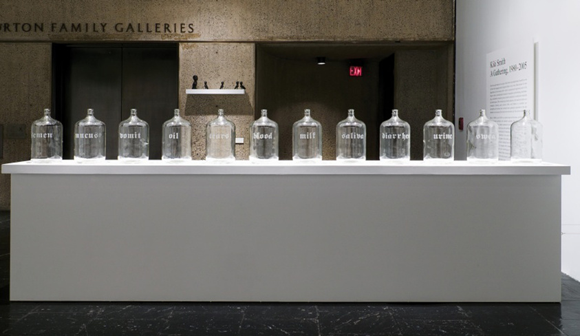Glass jars with various bodily fluids labeled on a white display in an art gallery.