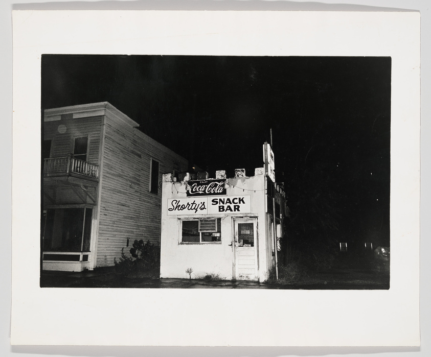 Black and white photograph of an old, two-story building at night with a sign reading "Shorty's SNACK BAR" and a Coca-Cola logo above the entrance. The building appears aged and the surrounding area is dark, suggesting the photo was taken in a rural or deserted location.