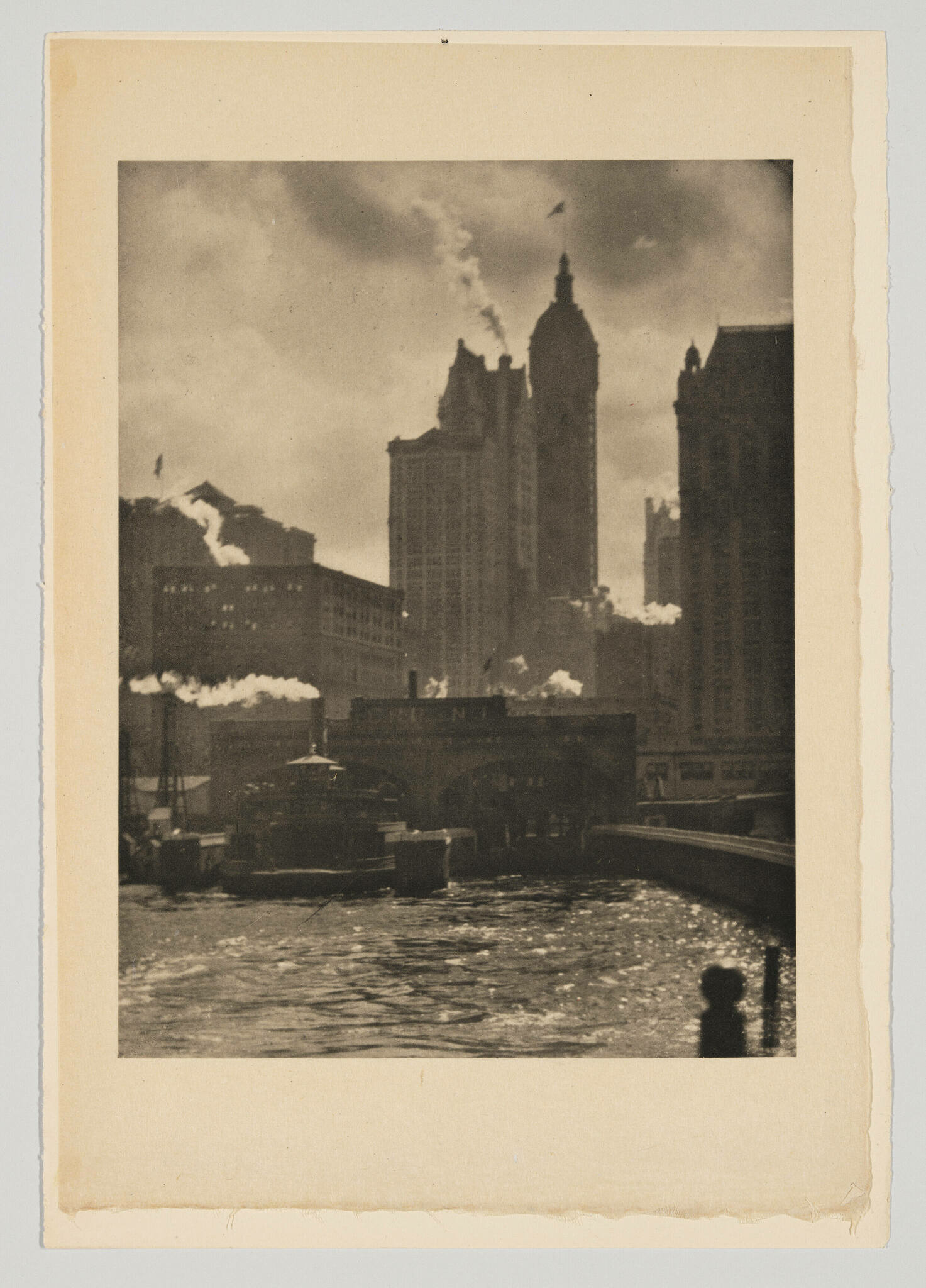 Vintage sepia-toned photograph depicting a cityscape with towering buildings, some emitting smoke from their rooftops, alongside a river with a boat. A bridge spans the river, and the silhouette of a person is visible in the foreground. The photo is mounted on a beige paper with rough edges.