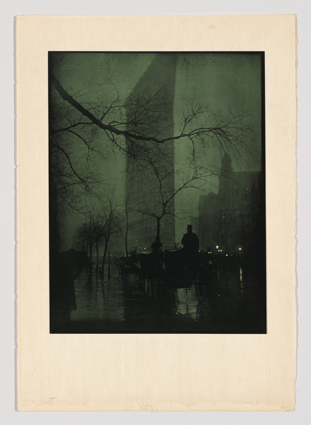 A vintage photograph depicting a nighttime street scene with silhouettes of bare trees, horse-drawn carriages, and a few pedestrians on a wet road, possibly after rain. The image has a dark, moody atmosphere and is mounted on a beige mat board with worn edges.
