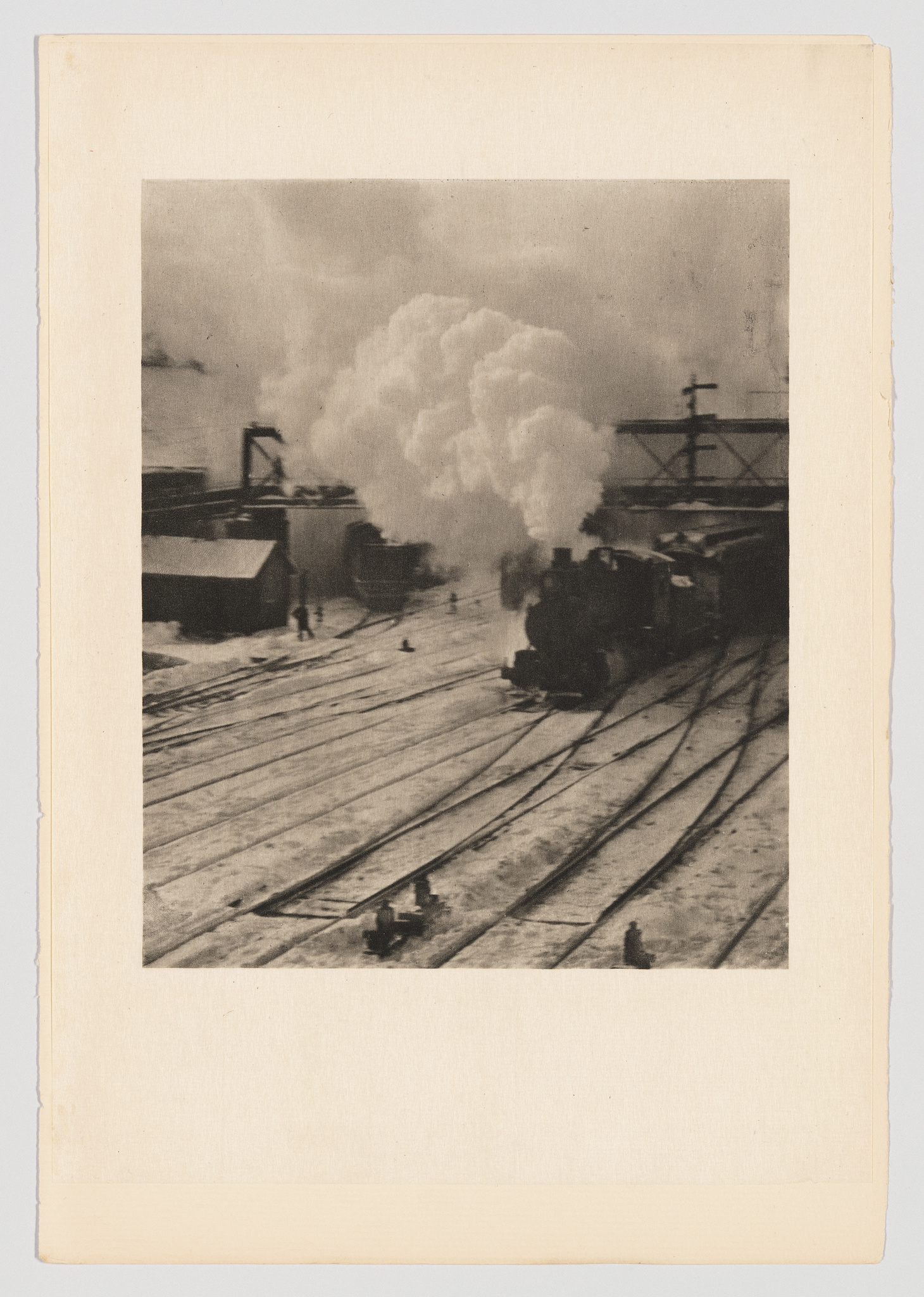 A vintage black and white photograph depicting a steam locomotive on railroad tracks, emitting a large plume of white smoke against an industrial backdrop with cranes and other structures. The image has a slightly worn appearance, with the edges of the photo mounted on a beige card.