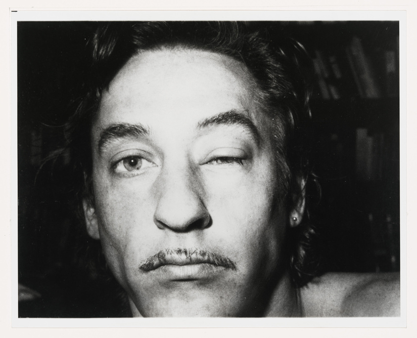 Black and white close-up photo of a man with a mustache, one eye closed, and a relaxed facial expression. The background is out of focus but appears to be a room with bookshelves.