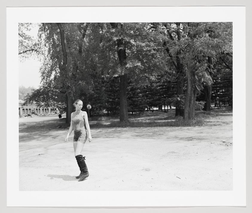 A black and white photograph of a woman walking across a park with tall trees in the background. She is wearing a sleeveless top, shorts, and knee-high boots. There is a building with arches visible in the far left background, and another person can be seen walking in the distance. The image has a border, suggesting it may be a print or framed.