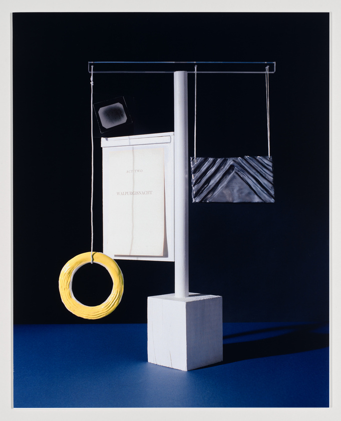 A conceptual art piece featuring a white vertical structure with a horizontal bar at the top from which various objects are suspended against a dark background. On the left, a black square object hangs above a white page with the text "ACT TWO WALPURGISNIGHT." In the center, a yellow circular ring dangles, and on the right, a metallic-looking folded fabric is suspended. The scene is set on a blue surface with a strong shadow cast by the white structure.